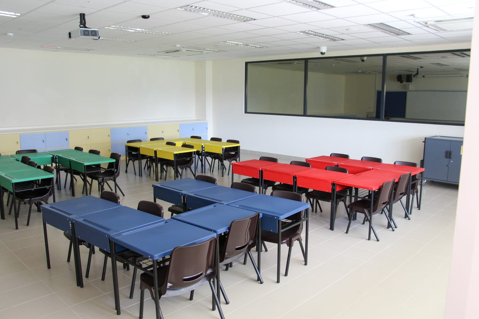 Empty classroom with groups of blue, red, yellow, and green tables surrounded by brown chairs, a projector on the ceiling.